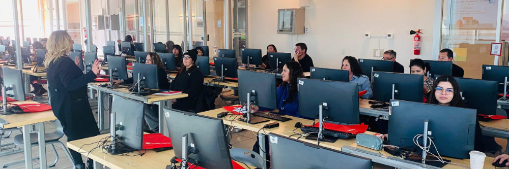 Estudiantes sentados en escritorio con computadores escuchando a la docente de pie